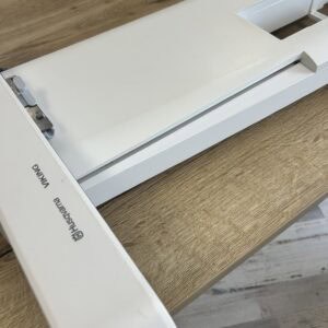Close-up of a white office device with a long horizontal slot on a wooden desk, likely a paper cutter/binding tool edge.