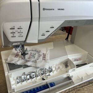 White Husqvarna Viking sewing machine with its open accessory case showing many presser feet and sewing tools laid out in trays.