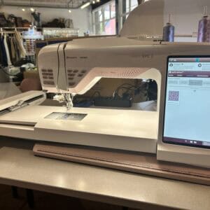 Designer EPIC 2 sewing machine by Husqvarna Viking on a sewing table in a fabric shop, with clothing racks and supplies in the background