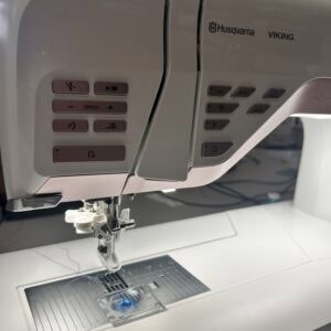 Close-up of a beige Husqvarna Viking sewing machine showing control panel buttons and the presser foot area, with the needle plate visible below.
