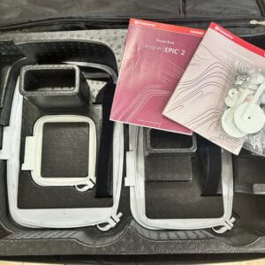 Open sewing machine case showing accessories, manuals, and small parts bags inside, including Husqvarna Viking Designer EPIC 2 guides.