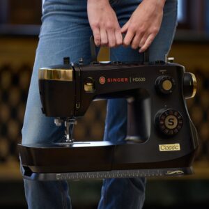 Person wearing jeans holds a black Singer HD500 sewing machine by its handle, viewed from the side.