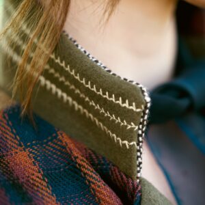 Close-up of an olive green wool collar with white blanket-stitch edging, worn over a blue and rust plaid garment.
