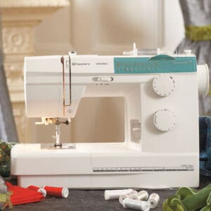 White sewing machine with turquoise control panel sits on a table, surrounded by loose thread spools and fabric scraps on the floor.
