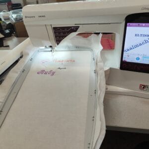 Close-up of a Husqvarna Viking embroidery machine stitching the word 'Ruby' with a pink heart on white fabric.