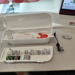 Open sewing kit with various threads, bobbins, and sewing accessories laid out on a table, next to a white sewing machine with a touchscreen panel.