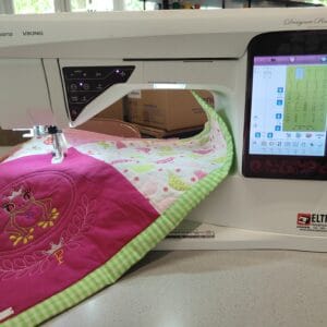 Front view of a Husqvarna Viking Designer Ruby sewing machine stitching a pink quilt with a green checkered border; touchscreen display visible on the right showing embroidery patterns.