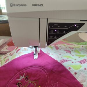 Sewing machine with 'Husqvarna VIKING' branding positioned over a colorful quilt with embroidered designs in pink and green threads.