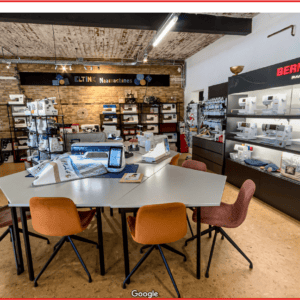 Bright sewing shop with rows of sewing machines, a central table, and orange chairs at the center of the room.