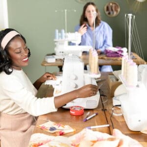 Two women sewing at industrial machines in a bright studio; the foreground woman smiles while guiding fabric, with sewing supplies on the table and a second seamstress in the background.