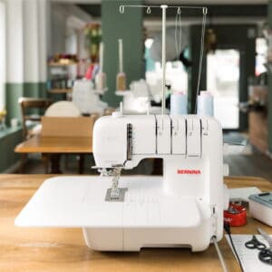 White Bernina sewing machine on a wooden table in a bright workshop, with spools of thread on top and equipment in the background.