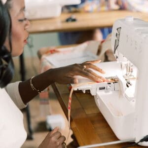 Close-up of a person guiding fabric through a white sewing machine at a wooden table.