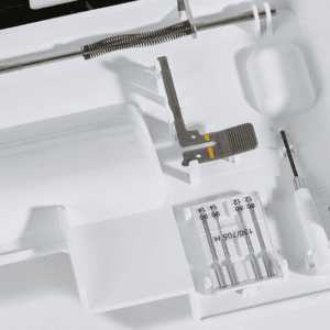 Close-up of a white sewing machine interior with tension discs, needle bar, and small screwdriver nearby.