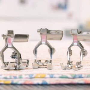 Three metal sewing machine presser feet lined up on a colorful quilt top, ready for stitching in a bright workspace.