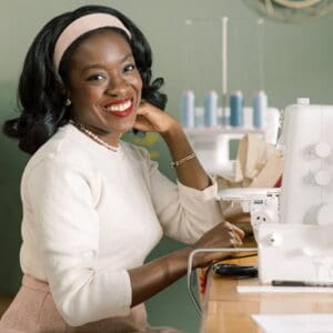 Smiling woman seated at a sewing machine in a bright workshop, wearing a cream sweater and headband.