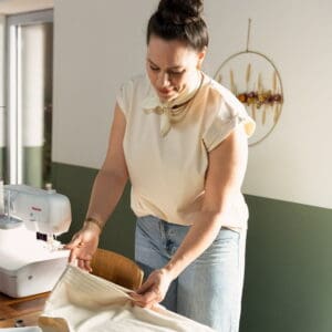 Woman in a light blouse and jeans sewing fabric with a white sewing machine on a wooden table, indoors.