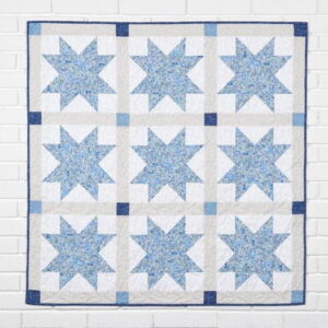 Quilt with a nine-star pattern in blue tones on white squares, bordered in navy, hanging on a white brick wall.