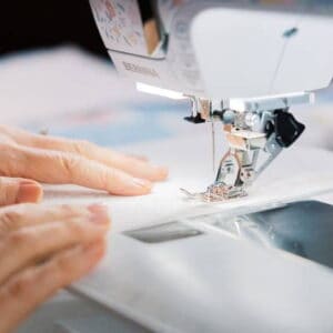 Person guiding fabric under a sewing machine needle, stitching precision on light-colored material.