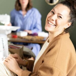 Woman smiling while sewing at a sewing machine in a bright workshop; another person works in the background.