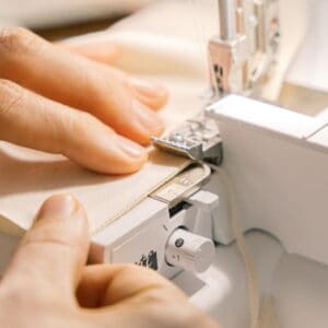 Close-up of hands guiding beige fabric under a sewing machine needle and presser foot while sewing an edge.