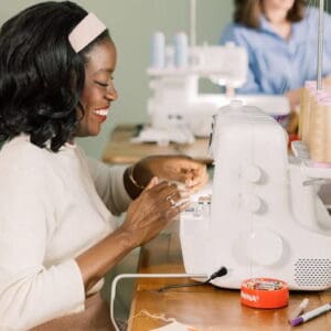 Smiling woman sewing on a white sewing machine at a wooden table, with thread and fabric nearby.