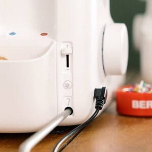 Close-up of a white Bernina sewing machine with the power cord plugged in and a red pin cushion on a wooden table.