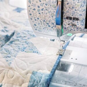A sewing machine stitching a blue-flower quilt with white and blue fabric panels beneath the needle area.