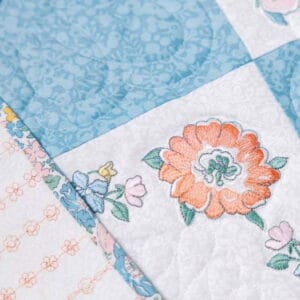 Patchwork quilt showing blue patterned squares and a white square with orange floral embroidery and pastel flowers.
