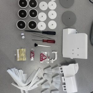 Assorted sewing machine parts on a table: black and white spool-like discs, gray round plates, white brackets, screwdrivers, tweezers, and fabric fringe on a gray surface.
