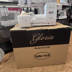 White Gloria Baby Lock sewing machine on its box, labeled Model BLETS8, in a store display with other machines on shelves in the background.