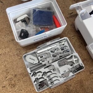 Top-down view of an open white toolkit with a blue cloth, plastic bags, and small hardware inside on a workshop floor visible beside another tool case with assorted bits and attachments.