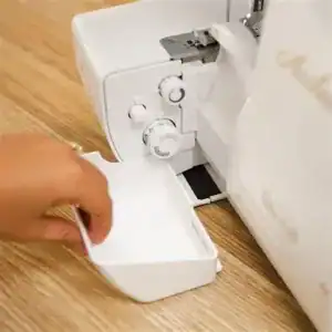 afvalopvangbak F1-70k01 - Eltink Naaimachines Close-up of a hand loading fabric into a small white accessory bin attached to a white sewing machine on a wooden table.