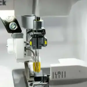 Close-up of an industrial sewing machine needle mechanism with metal parts and yellow tension indicators, shown in a neutral workshop setting.